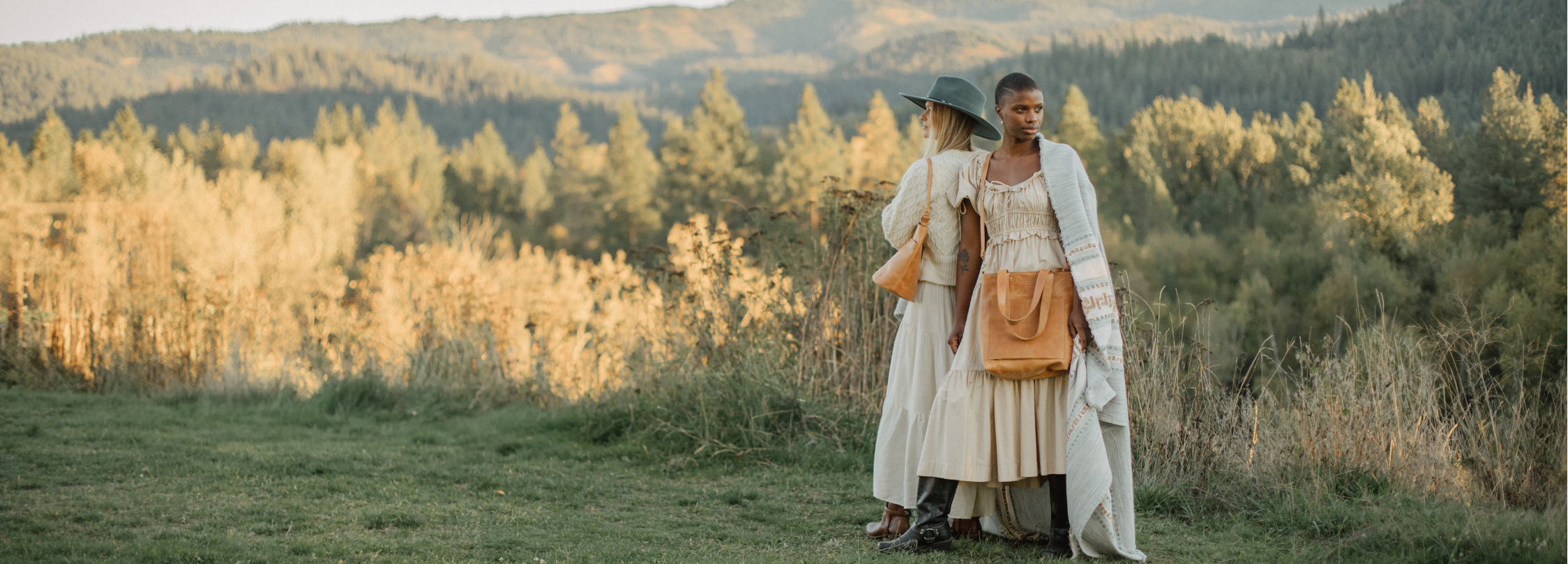 Two women in long white dresses and wide-brimmed hats stand outdoors in a grassy field with trees and hills in the background, holding tan crossbody bags.