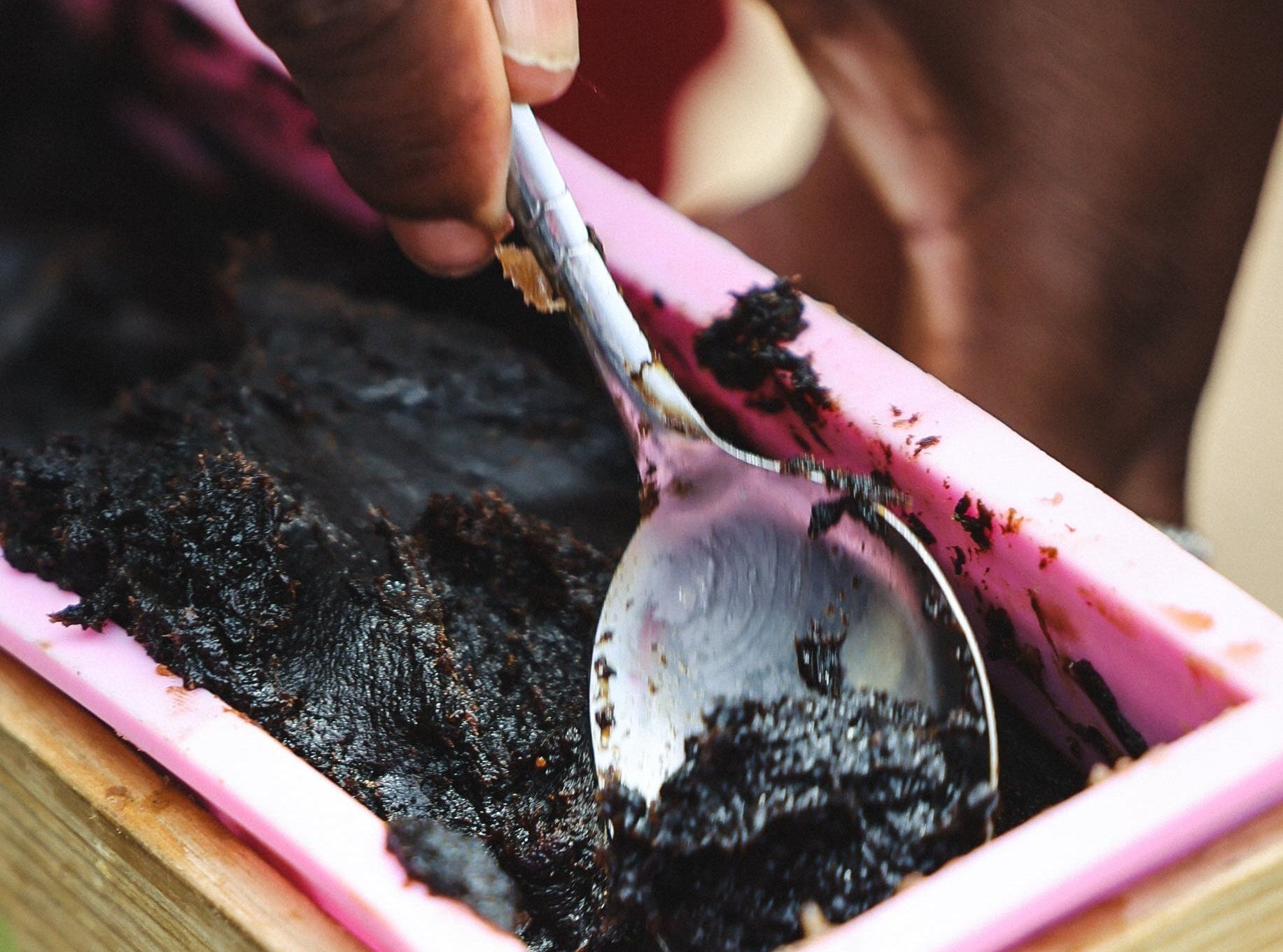 Person using a spoon to scoop dark substance from a pink container with a wooden base.