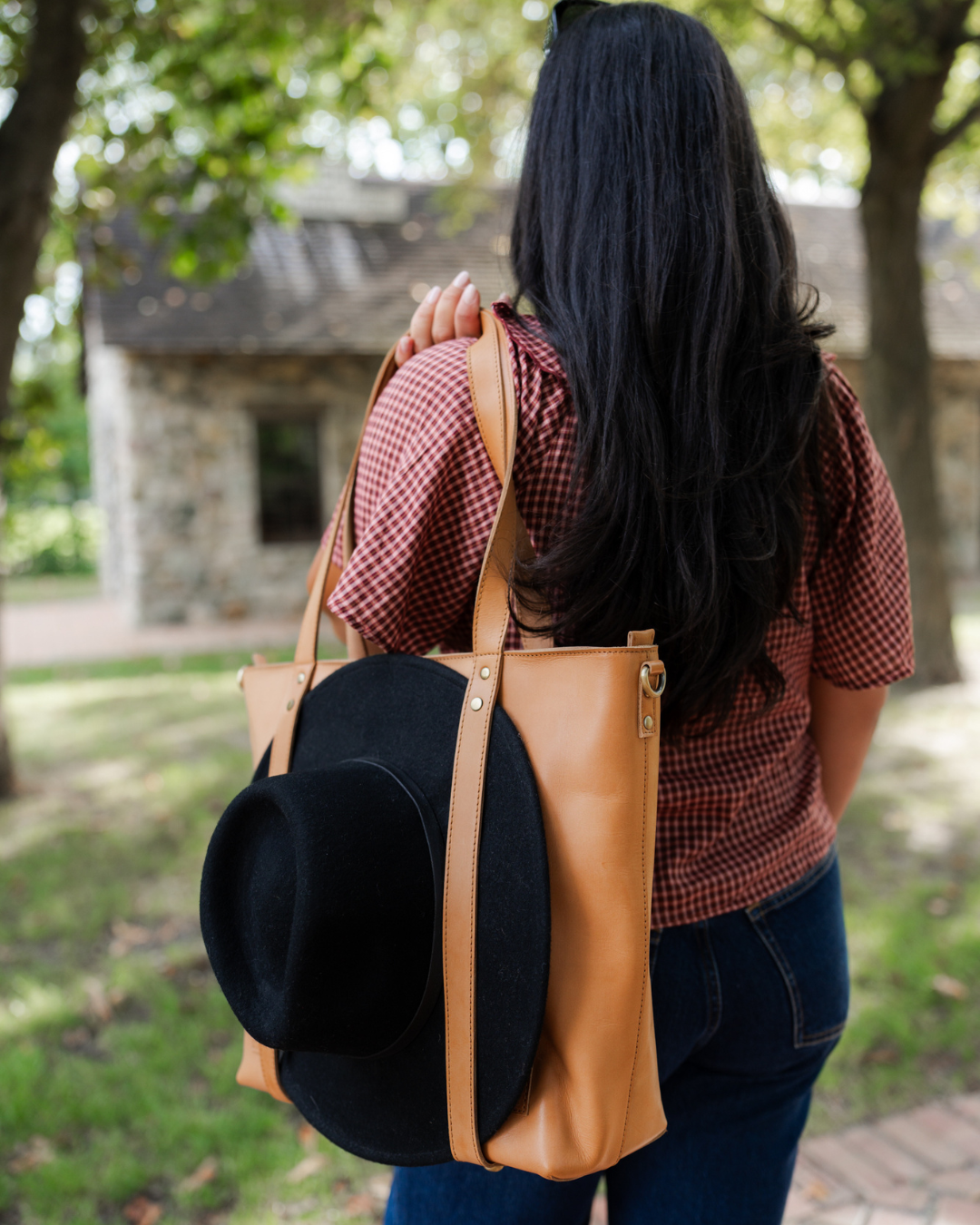 Person carrying a brown leather backpack with a black hat on top, outdoors.