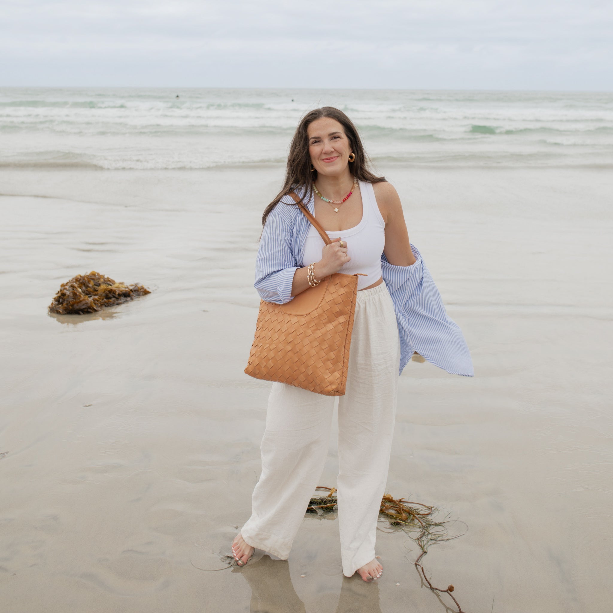 A woman standing by the beach, holding the Shirin Woven Leather Tote by Elevate, capturing the essence of casual elegance.