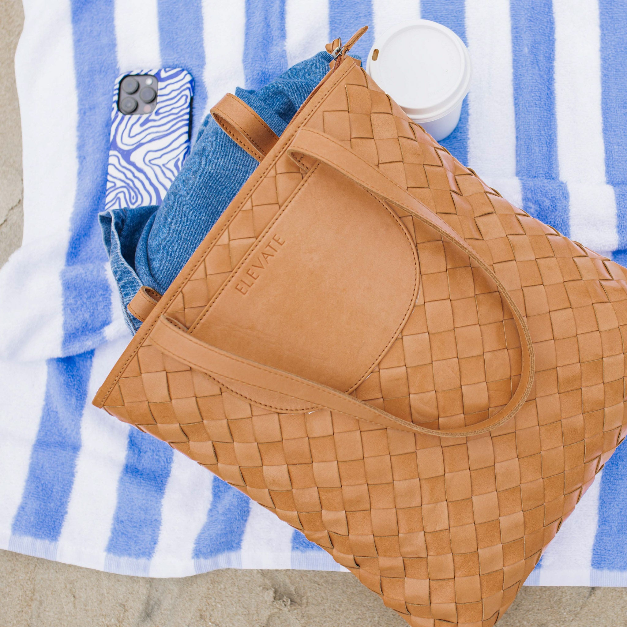 An overhead view of the Shirin Woven Leather Tote by Elevate, resting on a beach towel with denim and a coffee cup visible.