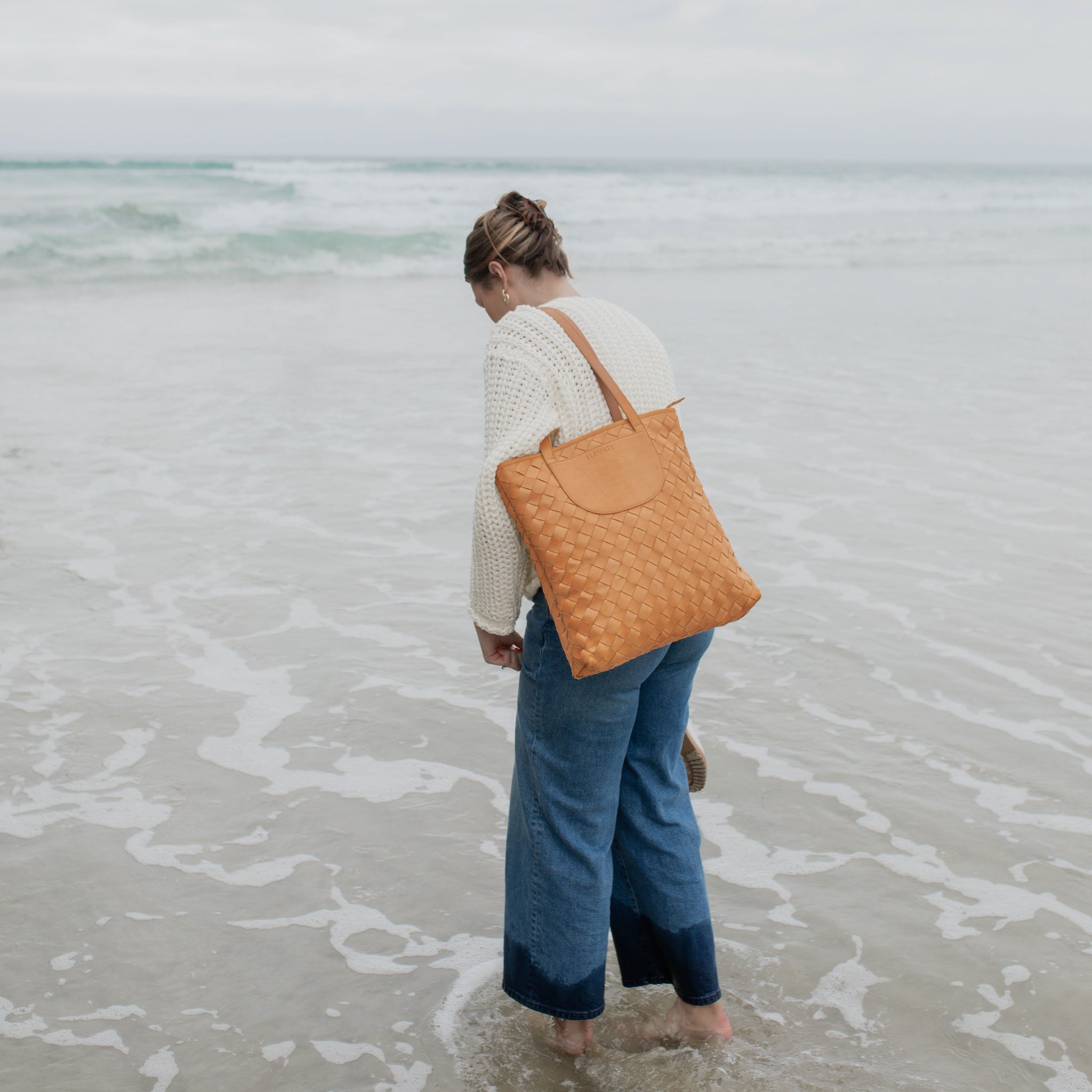 A model walking along the shore with the Shirin Woven Leather Tote by Elevate, emphasizing its beach-ready style.