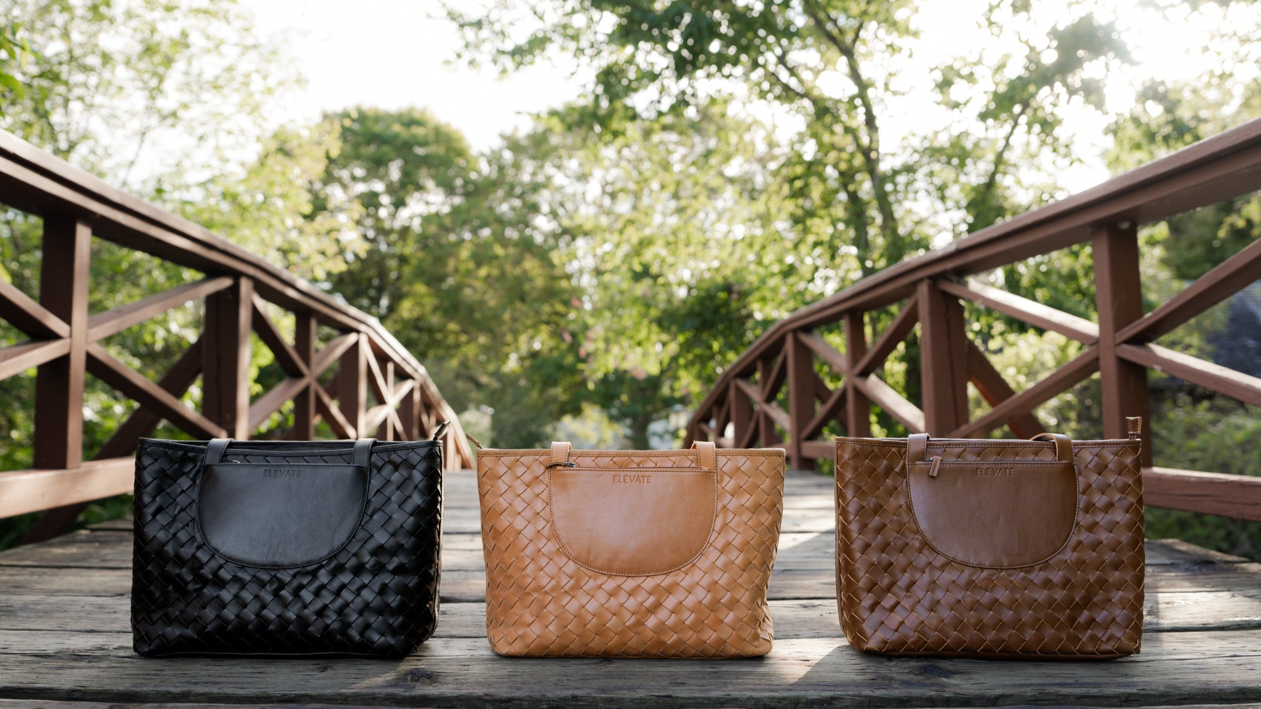 Three woven handbags on a wooden surface with a bridge and trees in the background