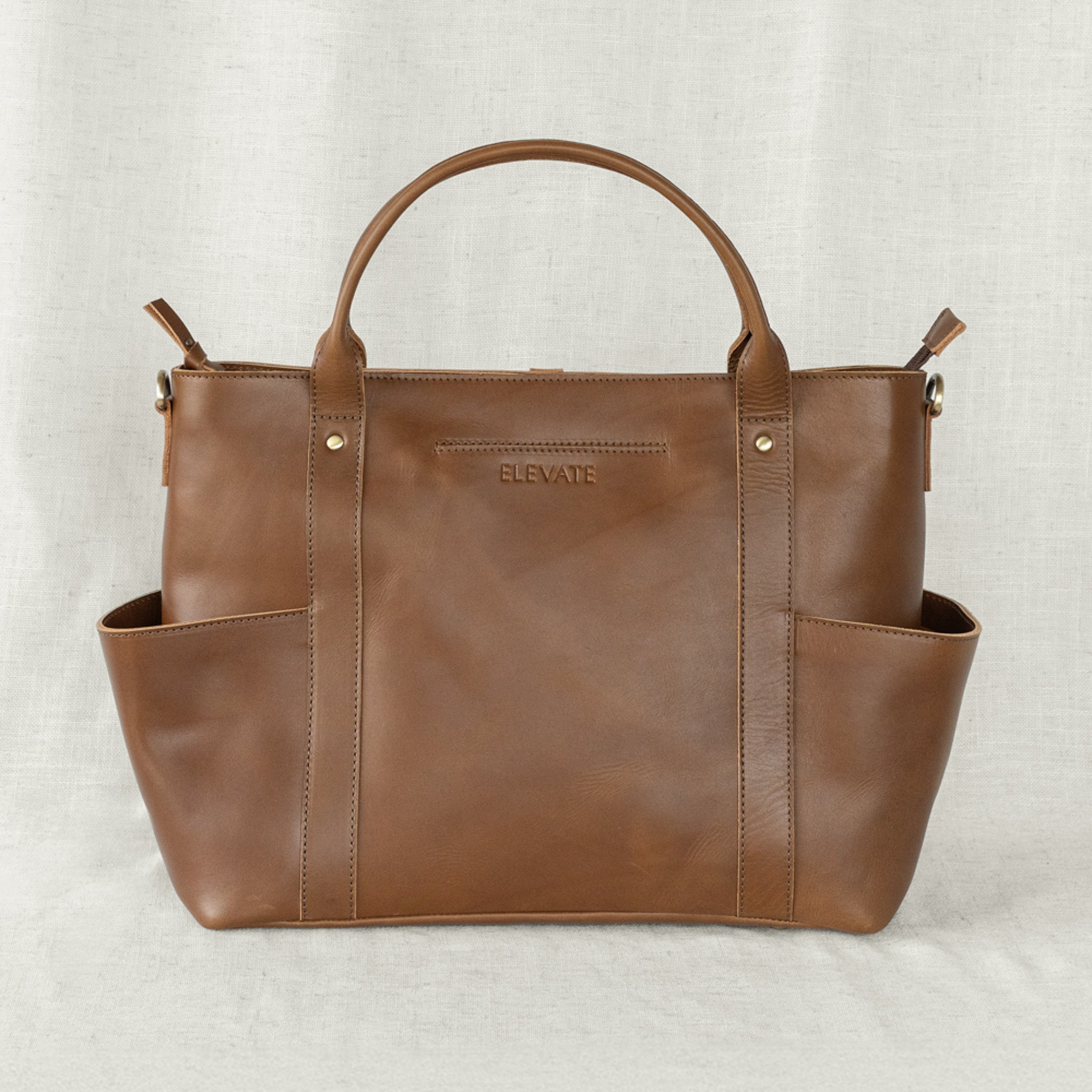 A chestnut leather utility bag with a structured design, visible brand name 'ELEVATE' on the front, and featuring a main compartment with additional external pockets.