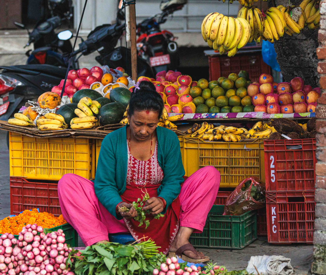 Woman sitting at a fruit and vegetable stand with various produce displayed.
