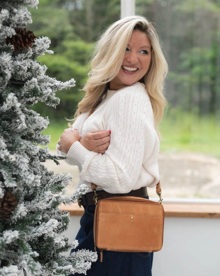 Woman in a white sweater holding a brown leather bag next to a snow-covered tree.