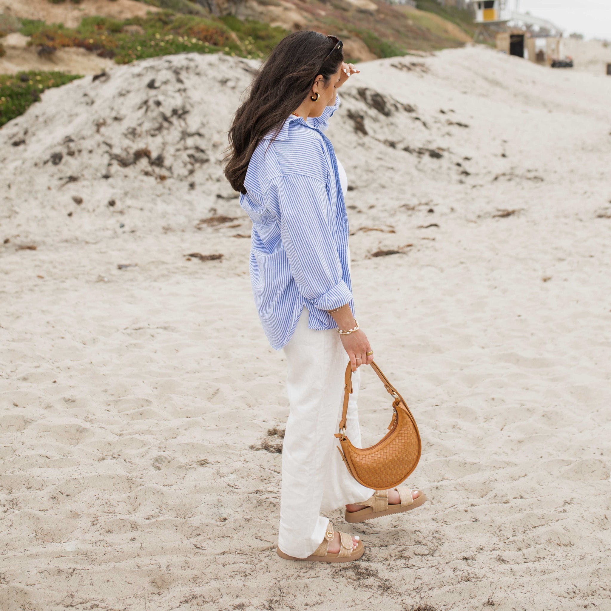 Woman walking on a sandy beach wearing a blue striped shirt and white pants, holding a brown handbag.