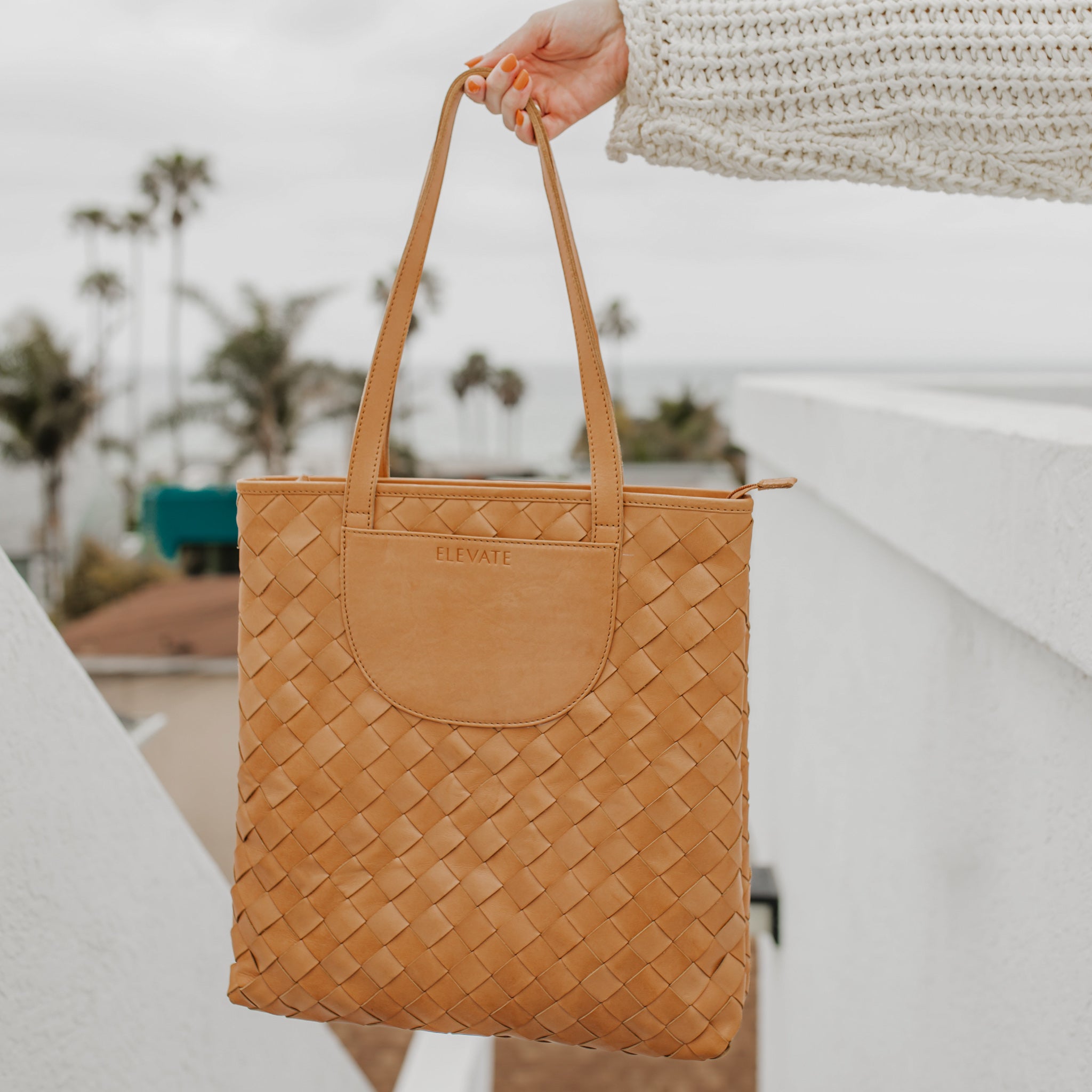 A stylish outdoor shot of the Shirin Woven Leather Tote by Elevate, held up against a coastal backdrop.