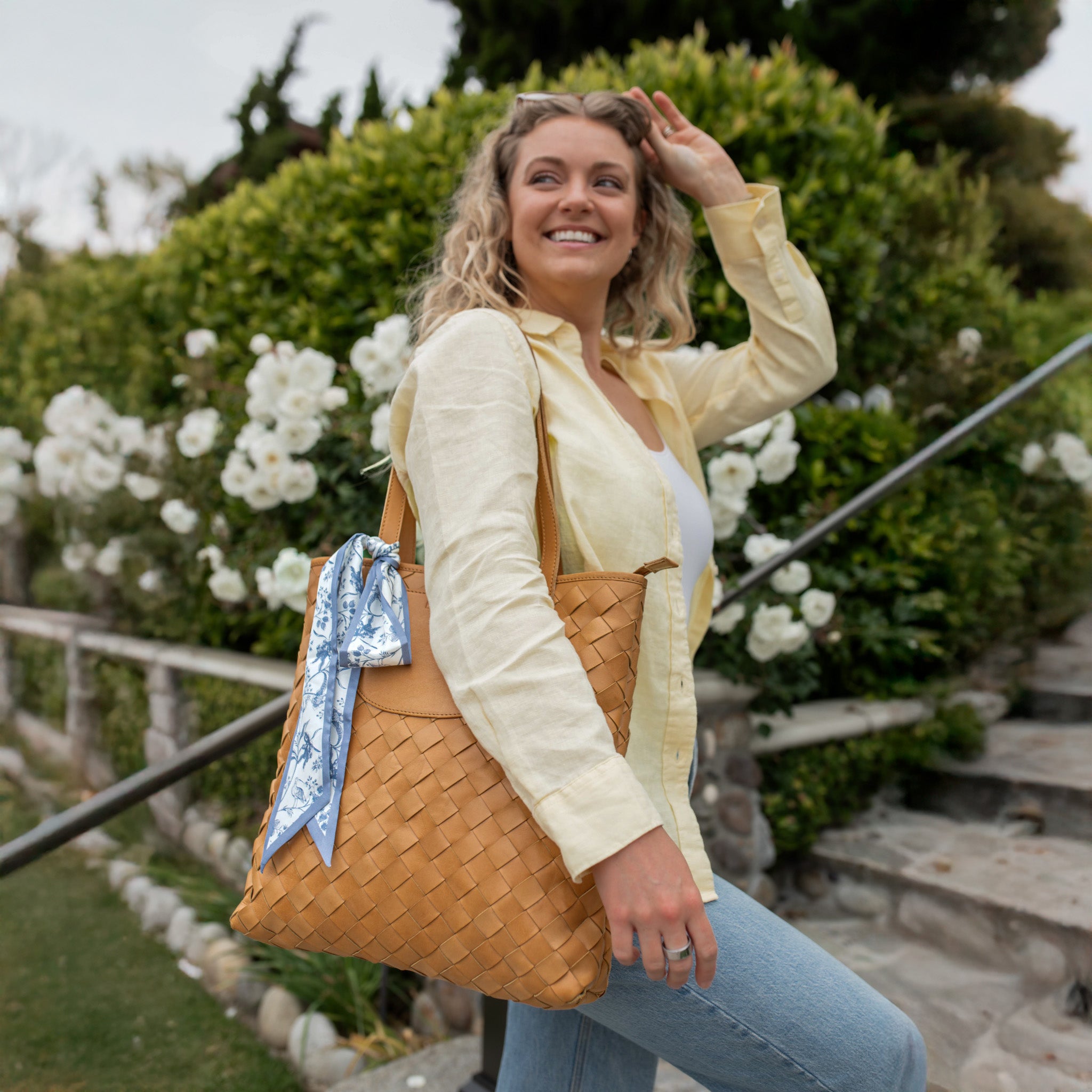 A model carrying the Shirin Woven Leather Tote by Elevate, showcasing the bag with a scarf and a playful smile in a garden setting.