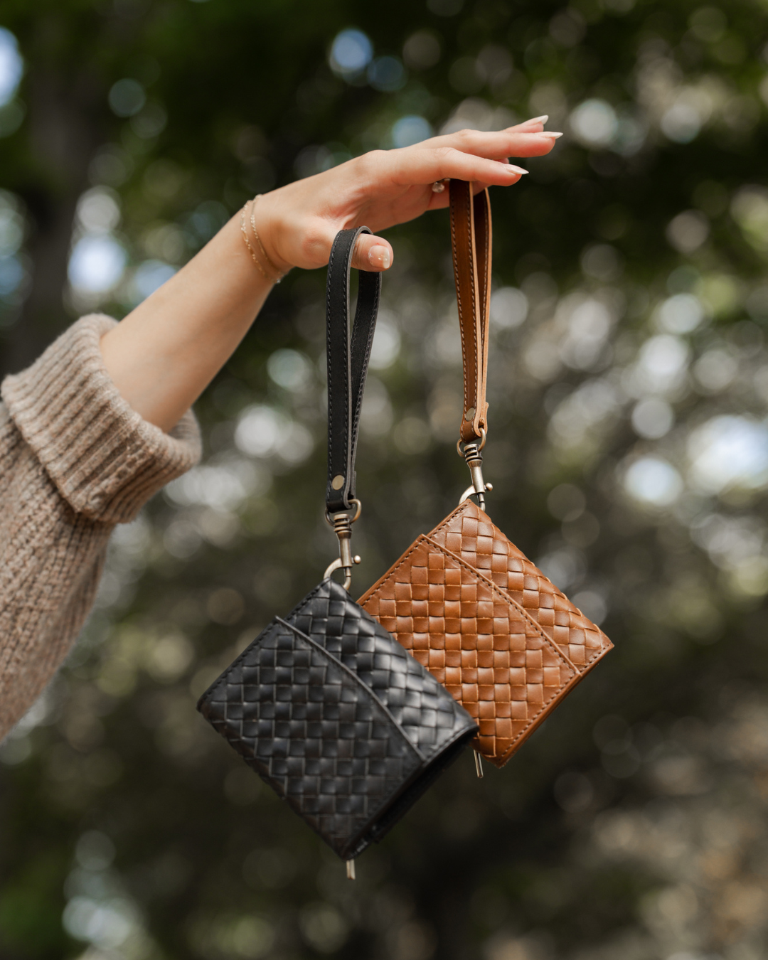 Hand holding two woven leather pouches, one black and one brown, against a blurred natural background.